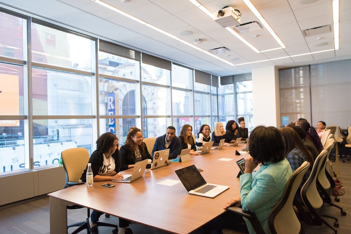 Group of colleagues seated around a long boardroom table with laptops in a glass-walled office